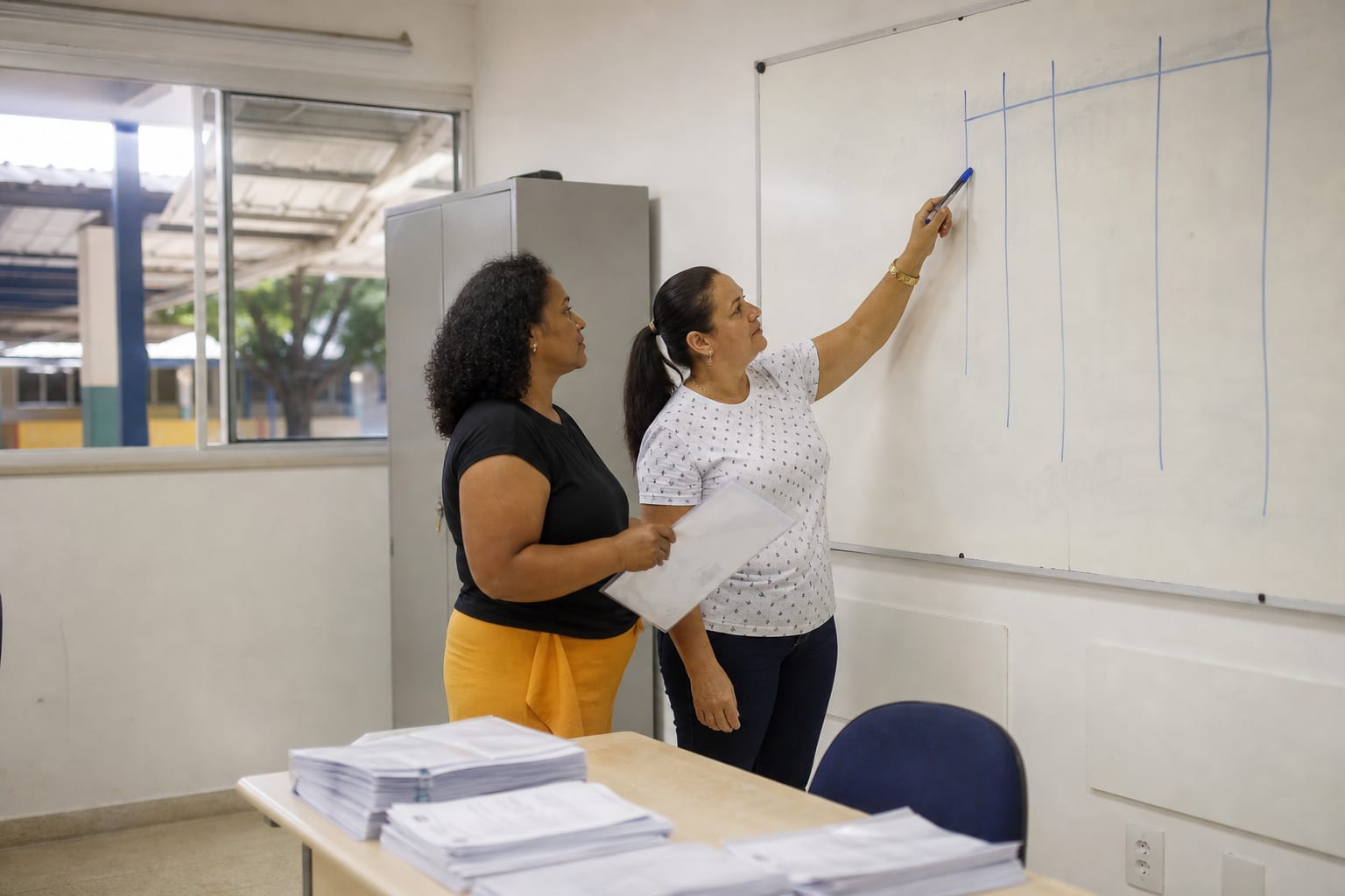 Duas profissionais de escola discutindo colunas desenhadas em um quadro branco em sala administrativa